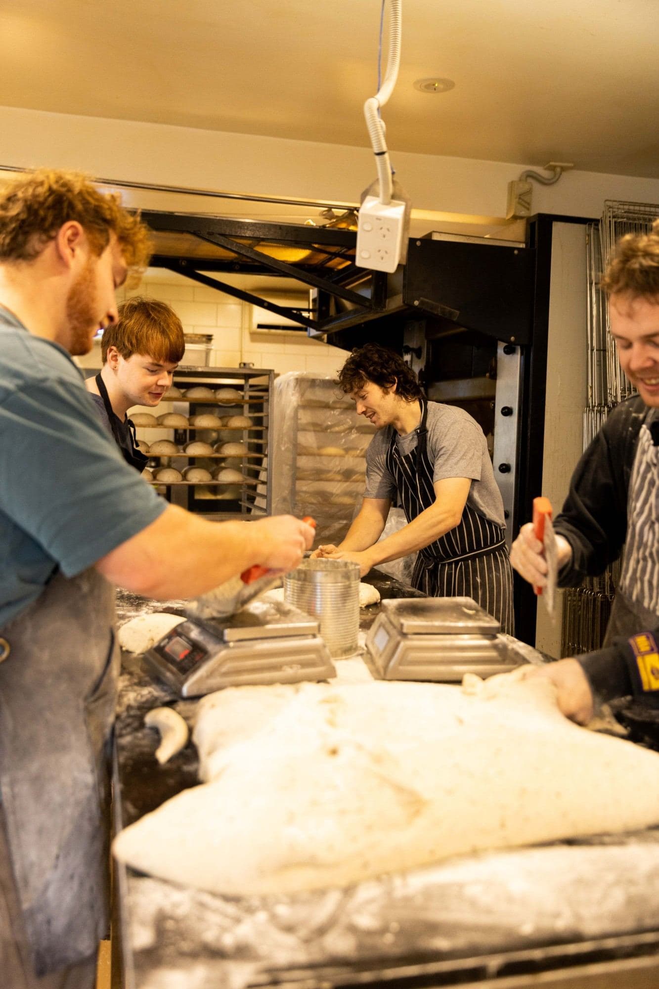 Four bakers dividing and shaping sourdough at the bench, with proving loaves on racks behind